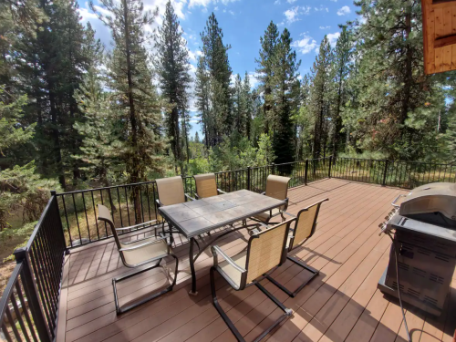 A spacious deck with a dining table and chairs, surrounded by tall trees and a clear blue sky.